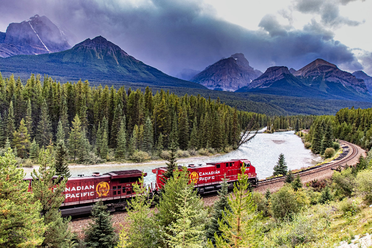 A Canadian Pacific freight train with two red locomotives curves through a lush coniferous forest alongside a turquoise glacial river, with dramatic, cloud-shrouded Rocky Mountain peaks rising in the background near Lake Louise, Alberta.