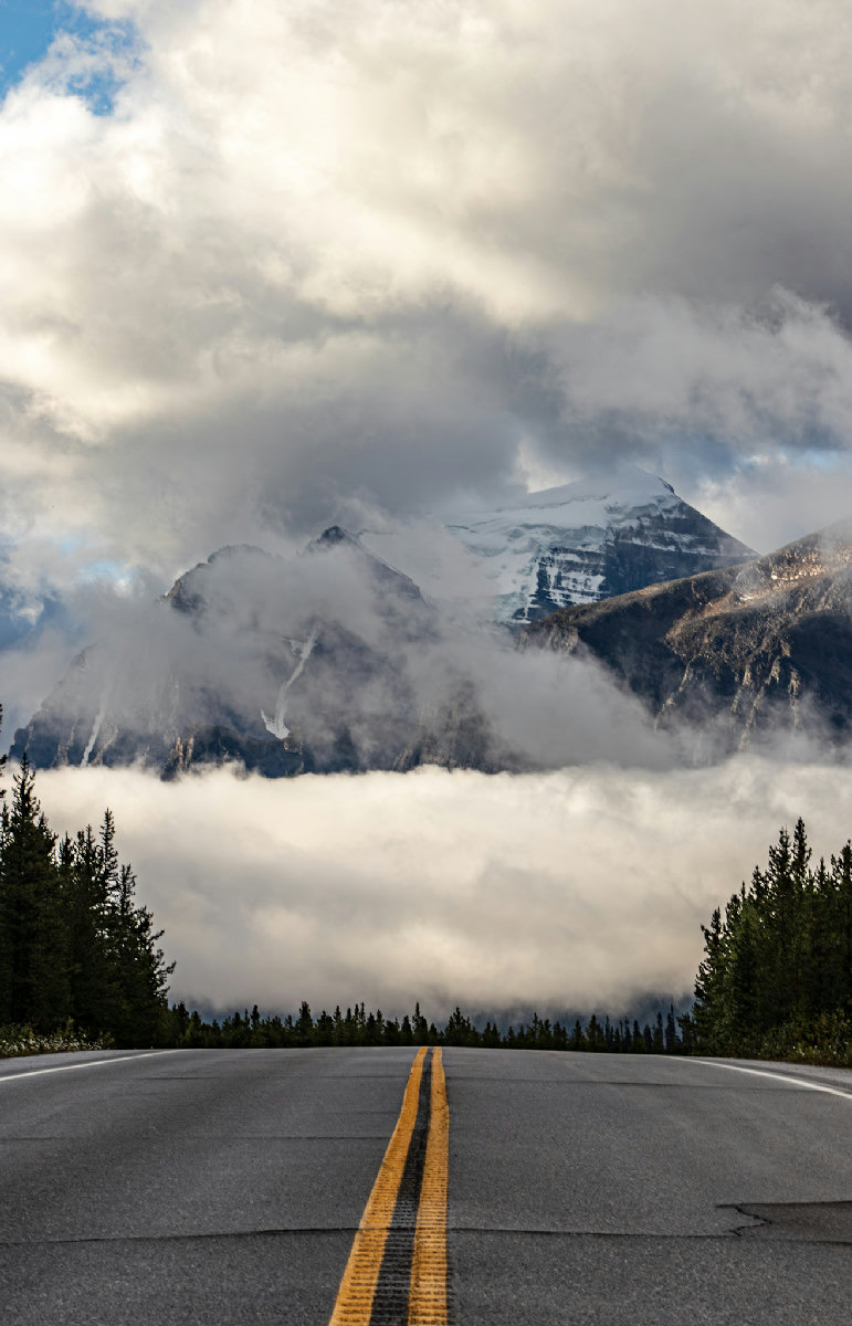 A two-lane asphalt highway with yellow centerlines stretches toward massive snow-capped mountain peaks dramatically shrouded in a low cloud inversion, with dark conifer forests framing both sides of the road under a brooding overcast sky.