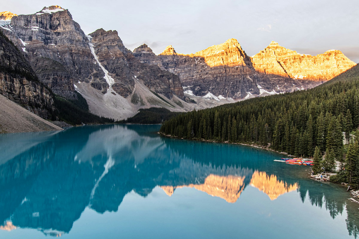 Moraine Lake at sunrise in Banff National Park, its brilliant turquoise glacial water perfectly reflecting the Valley of the Ten Peaks as warm alpenglow bathes the rocky summits in gold and orange. Dense evergreen forest lines the shore, and a small canoe dock is visible at the water's edge on the right.