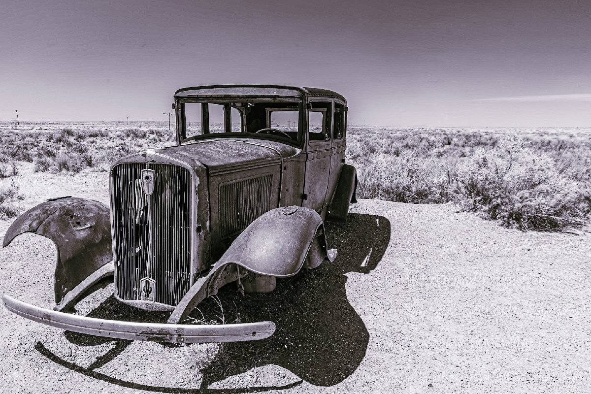 A rusted, deteriorating 1930s-era automobile sits abandoned on the desert roadside, its hood panels collapsed and chrome grille pitted with age, photographed in high-contrast monochrome with a wide-angle perspective that stretches the flat scrubland and pale sky to the horizon behind it.