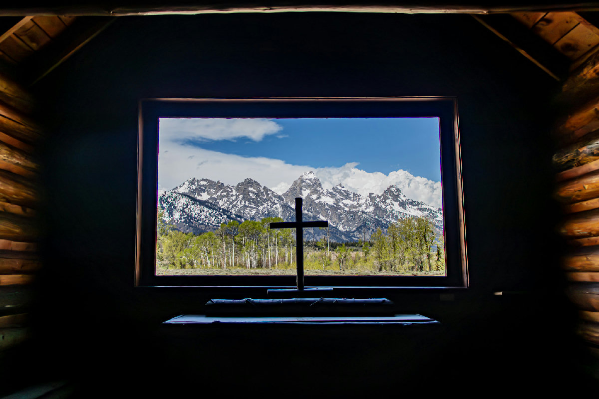 View from inside a dark log-walled chapel looking through a large rectangular window, where a wooden cross is silhouetted against a sweeping panorama of the snow-capped Grand Teton peaks under a bright blue sky with billowing clouds, with aspen groves glowing spring green in the valley below.